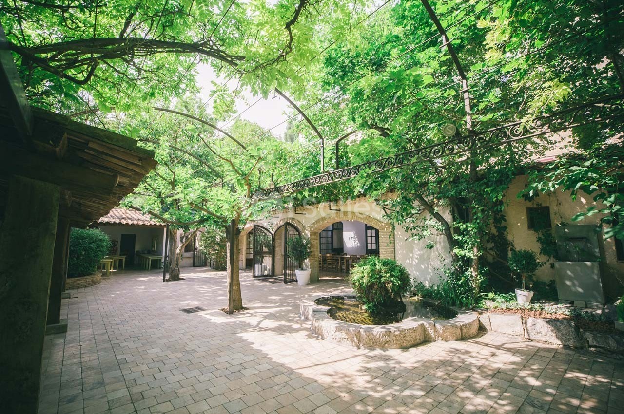 Cour intérieure pavée avec arbres et plantes grimpantes, entourée d'arches et d'une pergola en fer forgé.