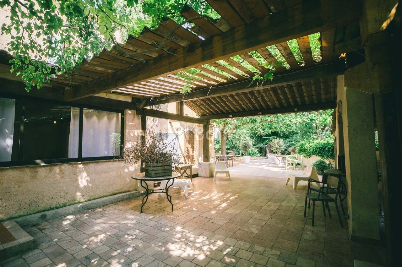 Pergola en bois ombragée sur une terrasse en pierre, entourée de végétation et de mobilier de jardin.