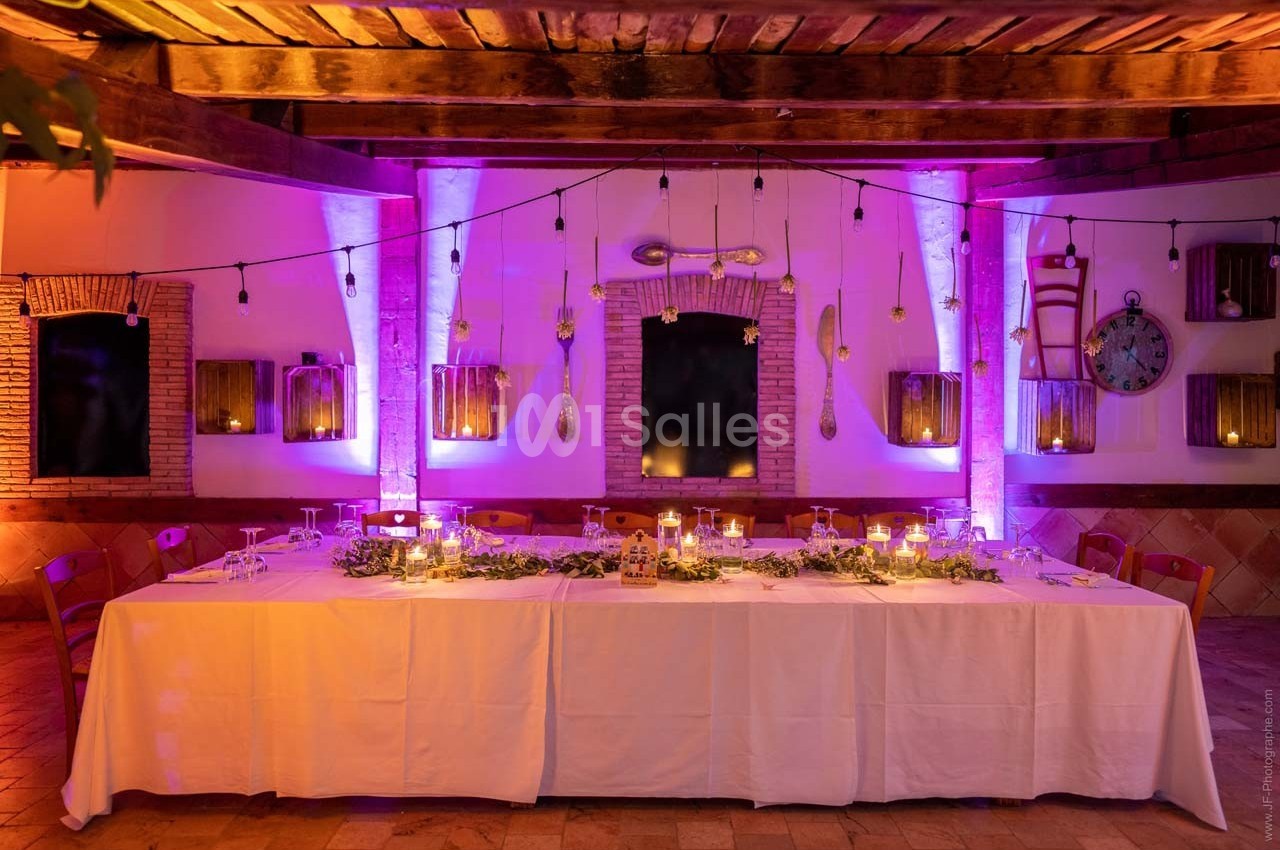 Table décorée pour un dîner, éclairée par des guirlandes lumineuses et des bougies, dans une salle aux murs en pierre.