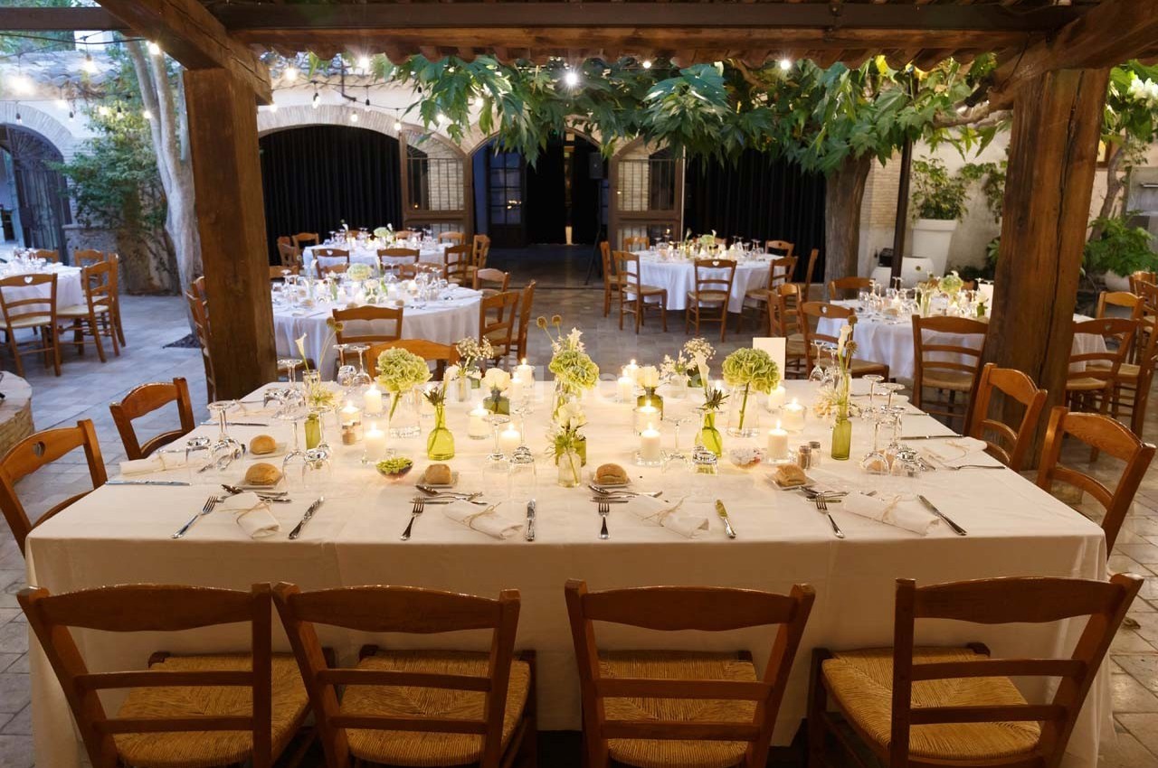 Grande table dressée pour un dîner en extérieur, entourée de chaises en bois, sous une pergola décorée de feuillage.
