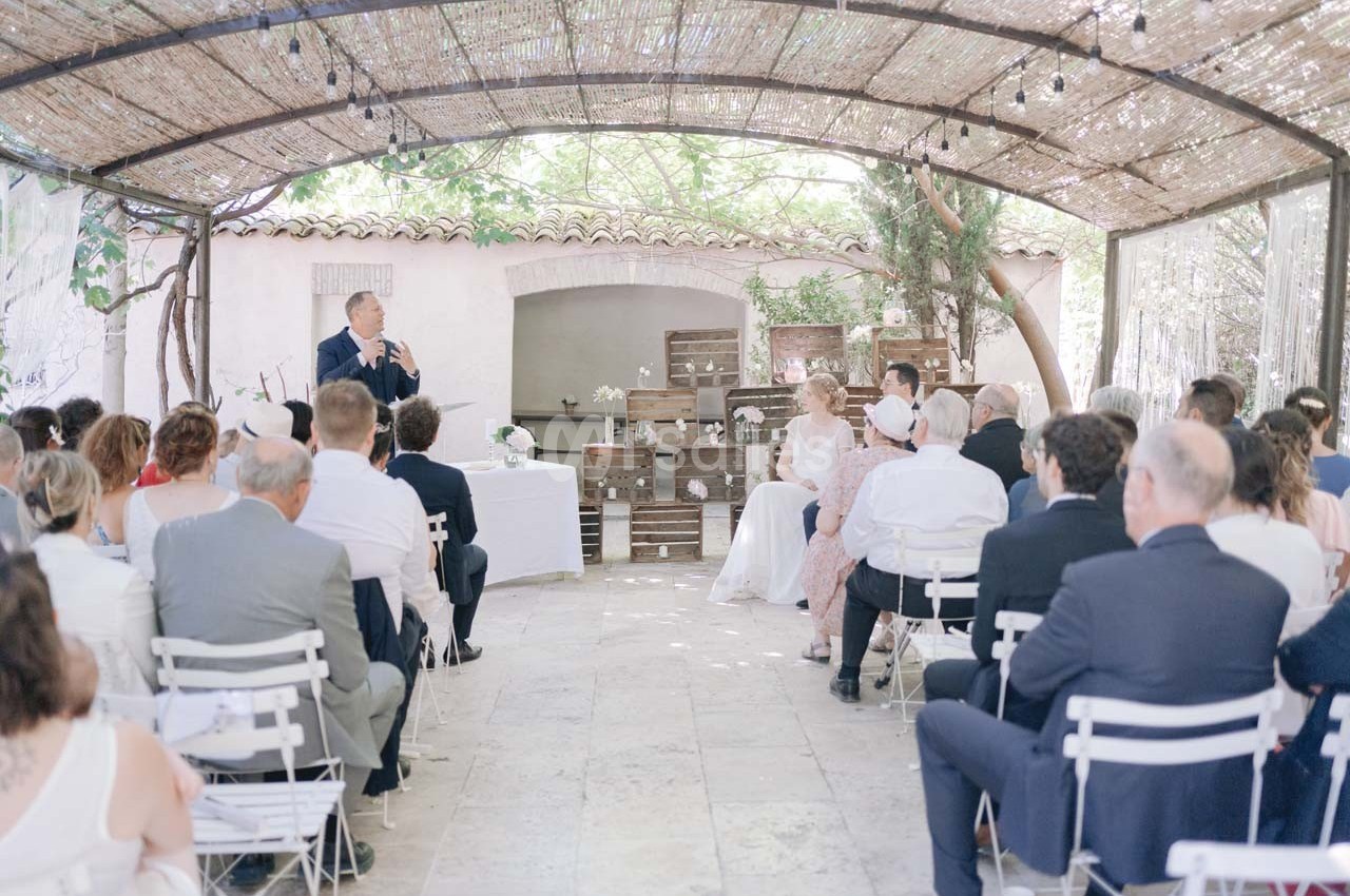 Cérémonie de mariage en extérieur sous une pergola, avec des invités assis et un orateur s'exprimant devant eux.