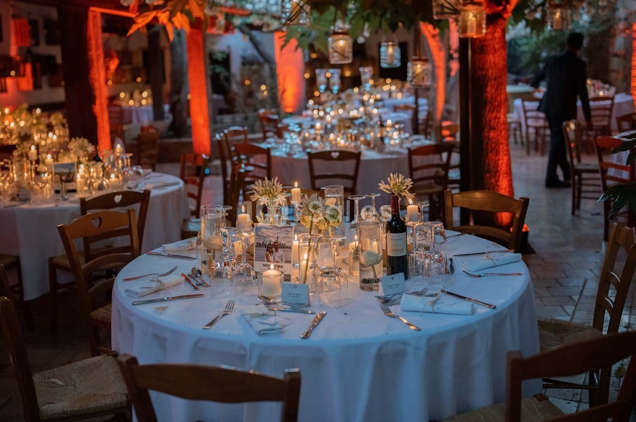Salle décorée pour un dîner, avec tables rondes dressées, bougies allumées et éclairage chaleureux.