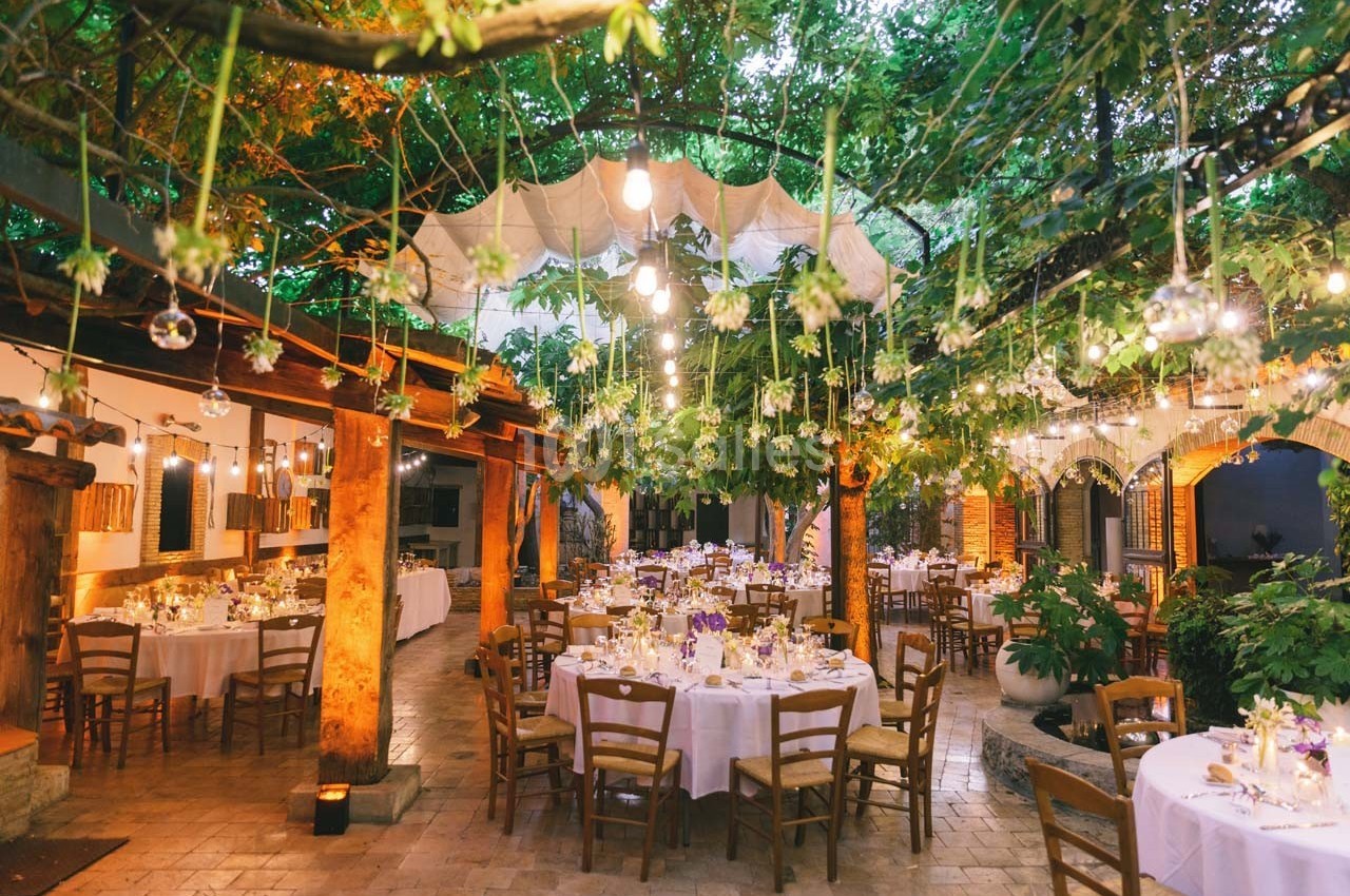 Salle de réception en plein air décorée de guirlandes lumineuses et de tables rondes dressées sous des arbres.