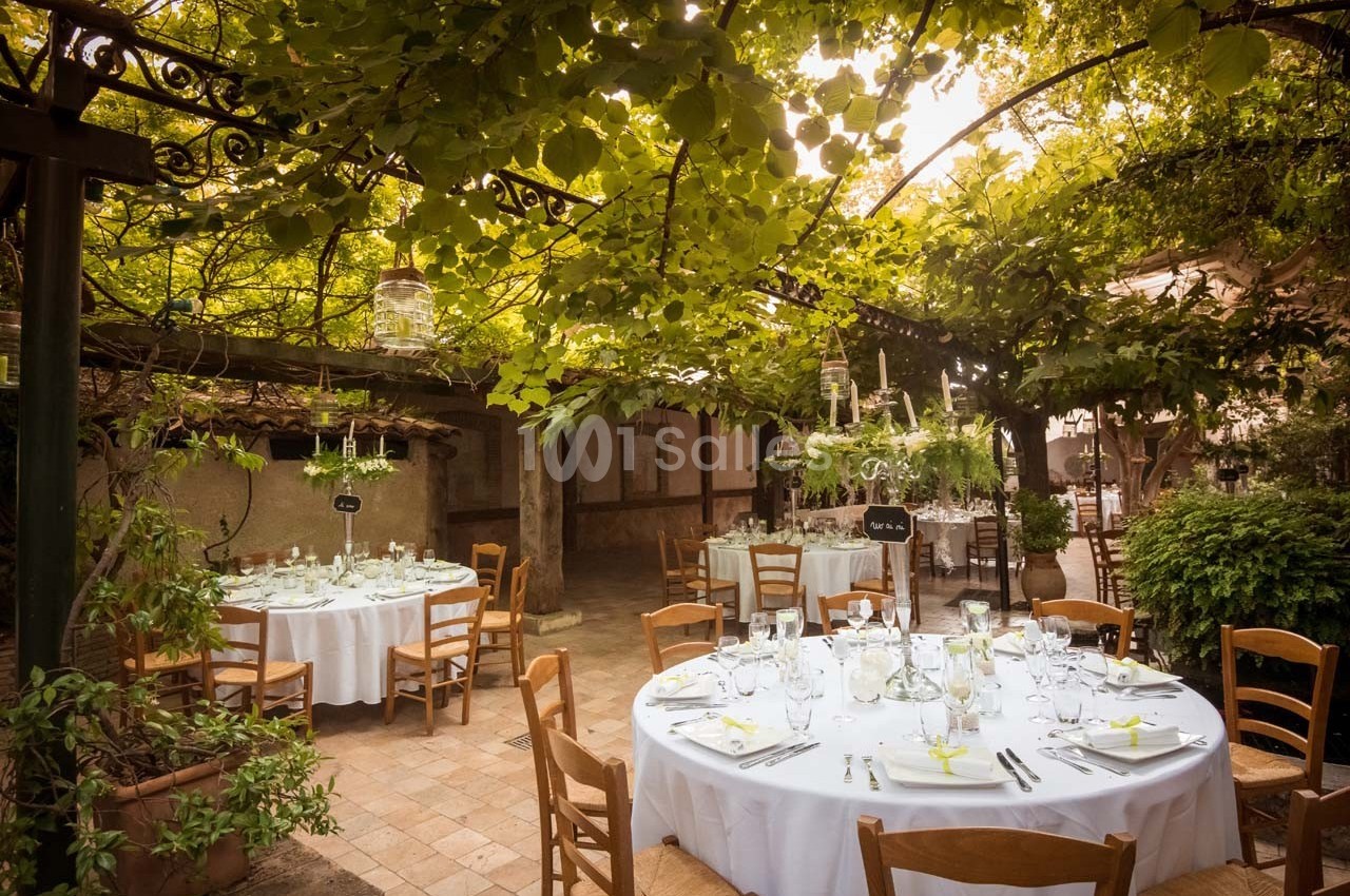 Tables rondes dressées avec nappes blanches sous une pergola couverte de feuillage dans un jardin ensoleillé.