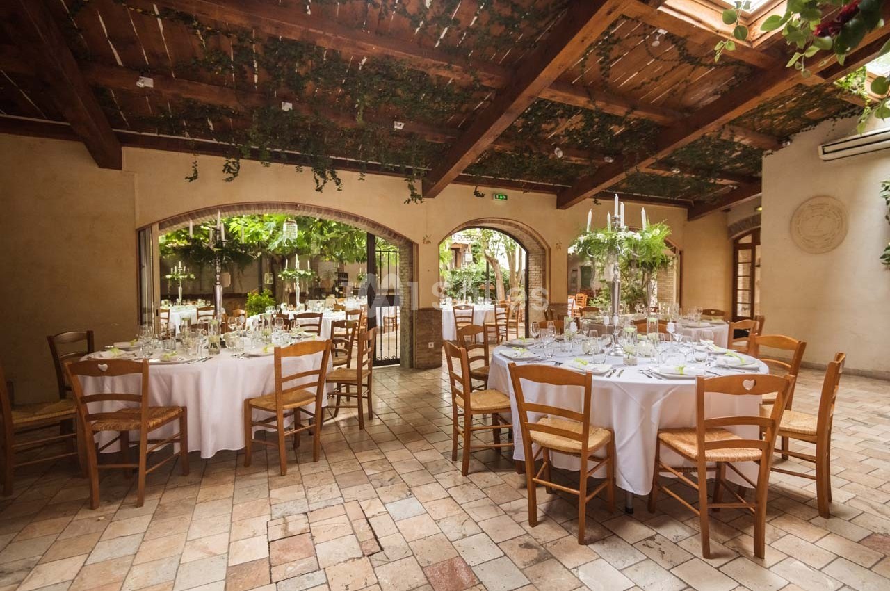 Salle de réception lumineuse avec tables rondes dressées, chaises en bois et plafond décoré de végétation suspendue.