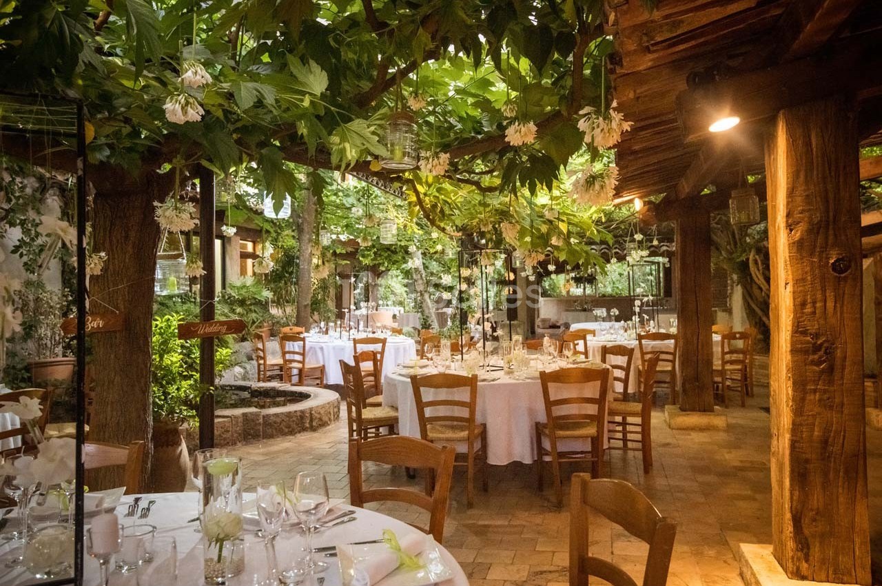 Salle de réception en plein air avec tables dressées, chaises en bois et végétation luxuriante sous un toit partiellement…