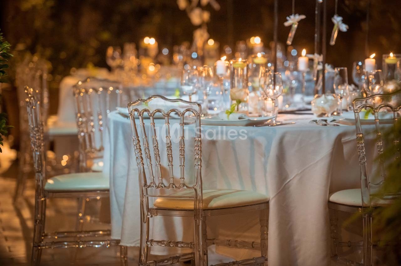 Table élégamment dressée avec nappes blanches, chaises transparentes et bougies allumées dans une ambiance tamisée.