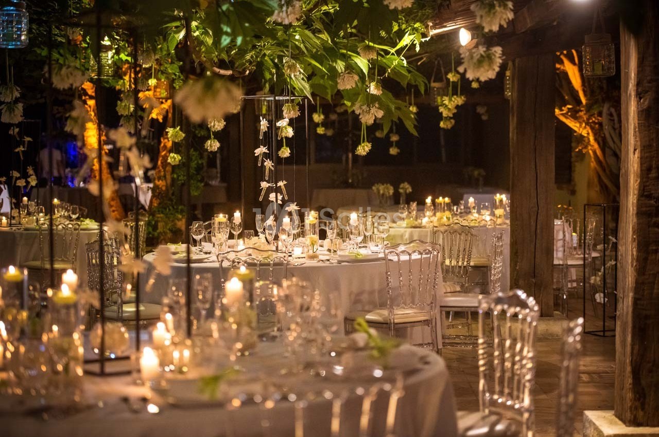 Salle de réception décorée de guirlandes lumineuses, bougies et fleurs suspendues, avec tables élégamment dressées.