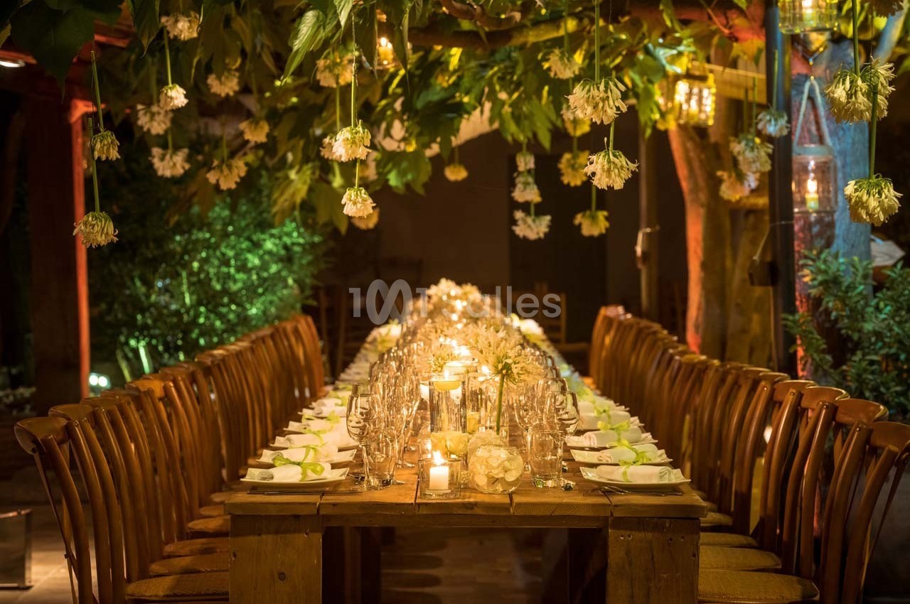 Table en bois décorée pour un dîner, entourée de chaises, avec éclairage chaleureux et fleurs suspendues.