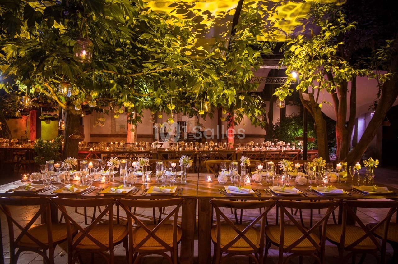 Table dressée pour un dîner en extérieur, entourée d'arbres illuminés et décorée de bougies et fleurs.