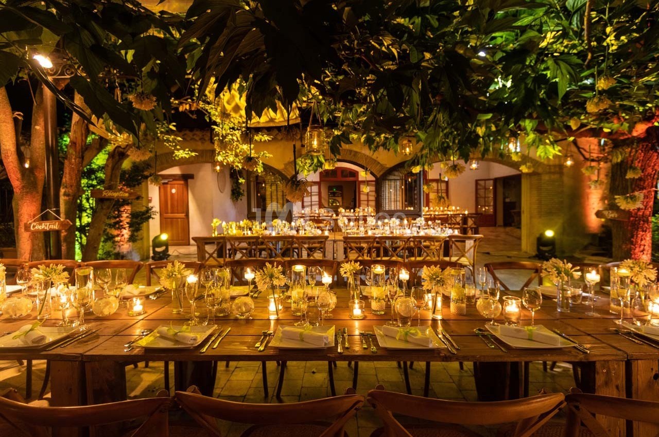 Tables en bois décorées de bougies et de fleurs jaunes, disposées dans un jardin éclairé pour un dîner en soirée.