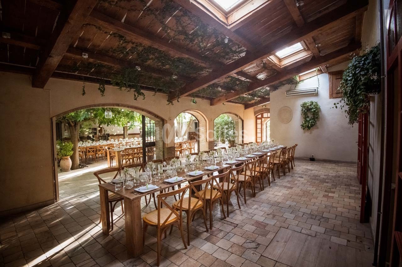 Salle lumineuse avec tables en bois dressées pour un repas, grandes baies vitrées et plantes suspendues.