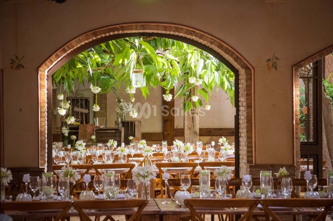 Salle de réception décorée avec des tables en bois, des fleurs blanches et une vue sur un arbre à travers une arche en…