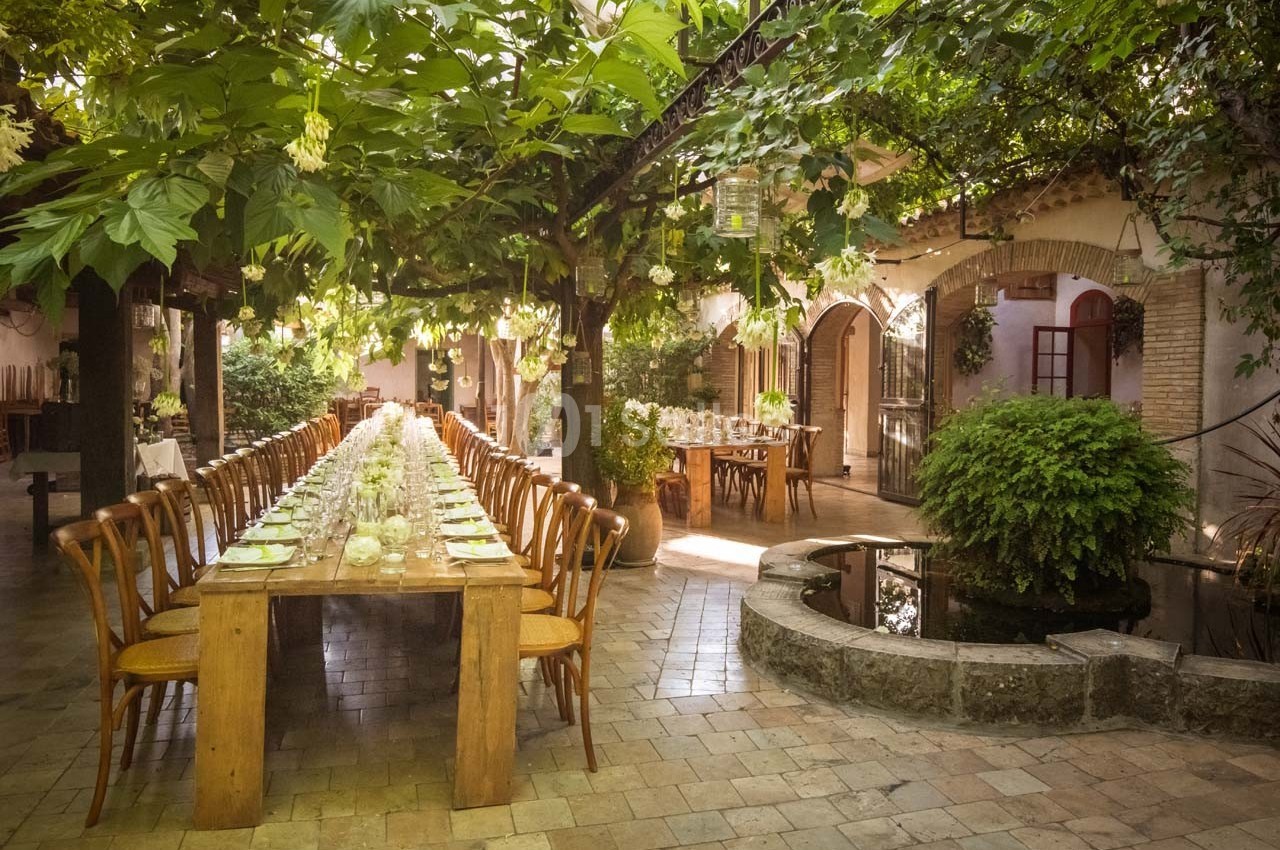 Grande table en bois dressée sous une pergola verdoyante dans une cour pavée avec un petit bassin.