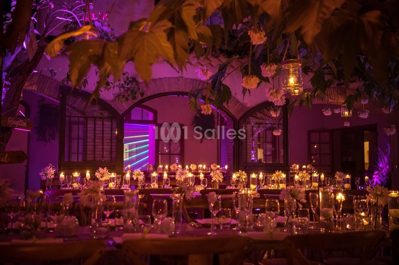 Salle décorée pour un dîner, éclairée par des bougies et des lumières tamisées, avec des tables ornées de fleurs.