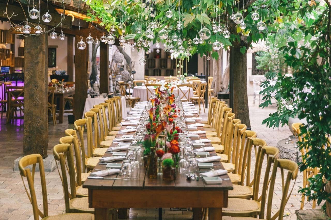 Grande table en bois décorée de fleurs colorées, entourée de chaises, sous un toit végétalisé avec des lumières suspendues.