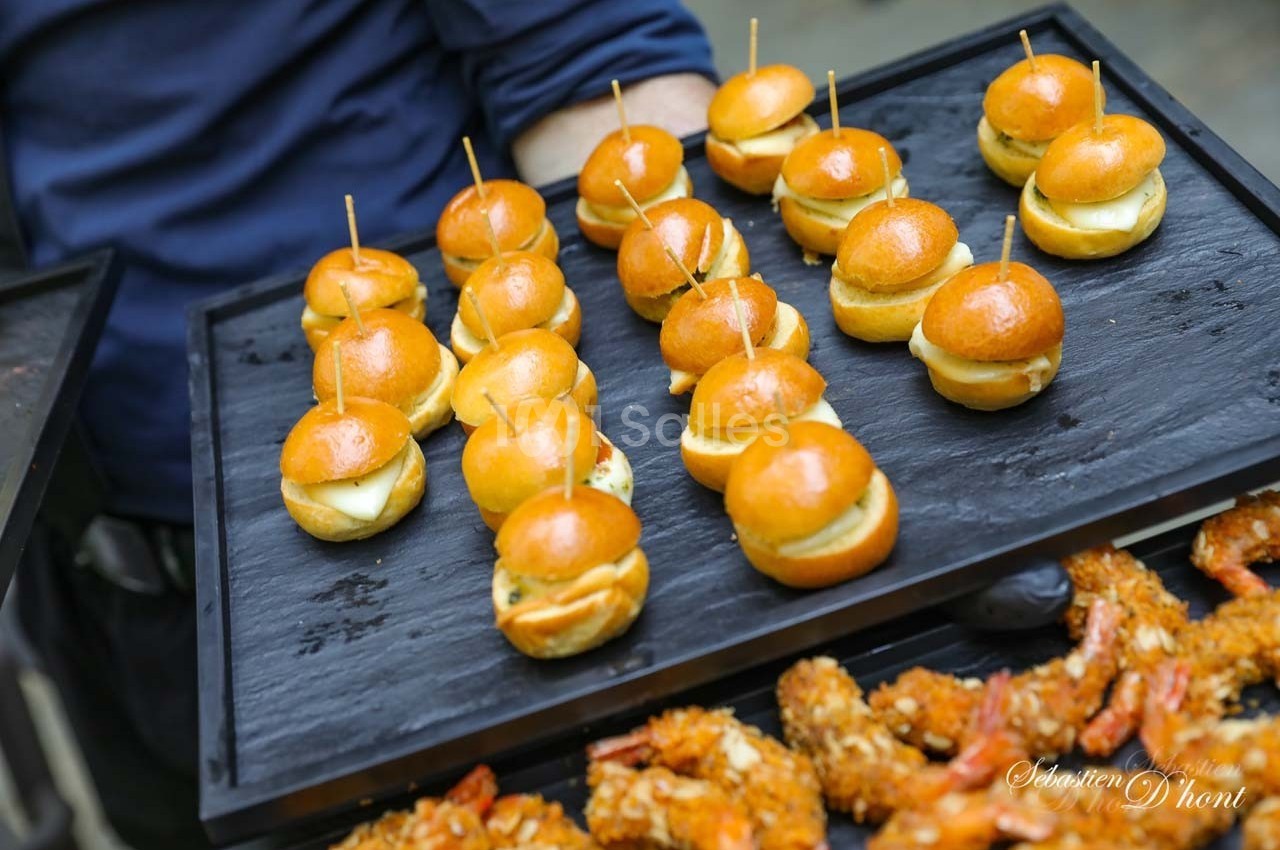 Plateau de mini-burgers dorés et brochettes de crevettes panées disposés sur un support noir.