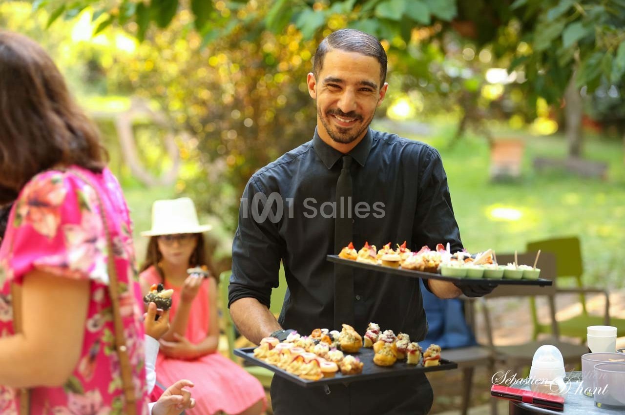 Un serveur souriant présente des plateaux de bouchées apéritives dans un jardin lors d'un événement en plein air.