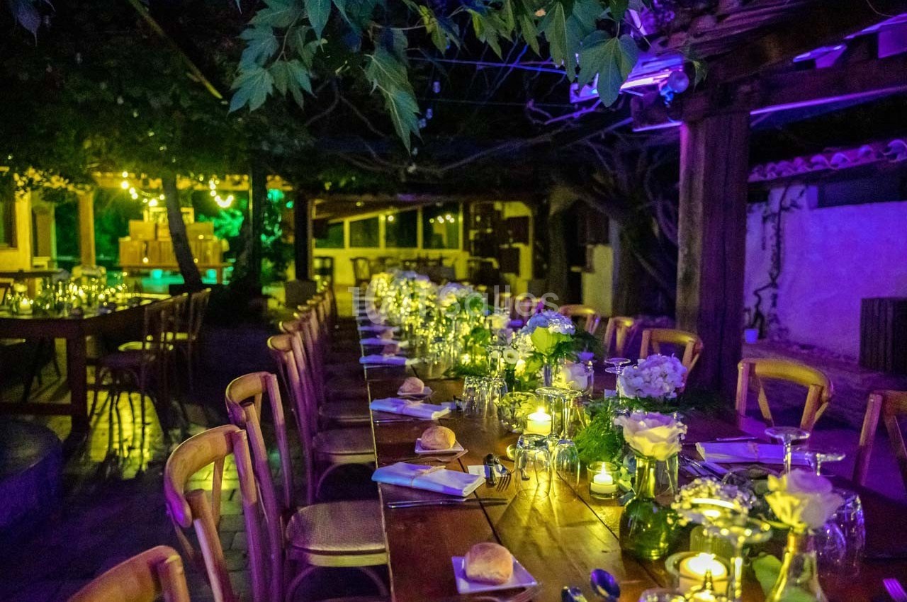 Grande table en bois décorée de fleurs et bougies, installée en extérieur sous des lumières tamisées.
