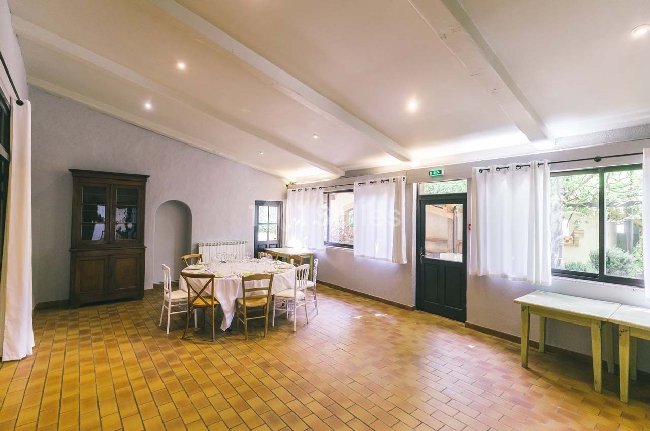 Salle lumineuse avec carrelage en terre cuite, tables dressées, chaises blanches et grandes fenêtres avec rideaux.