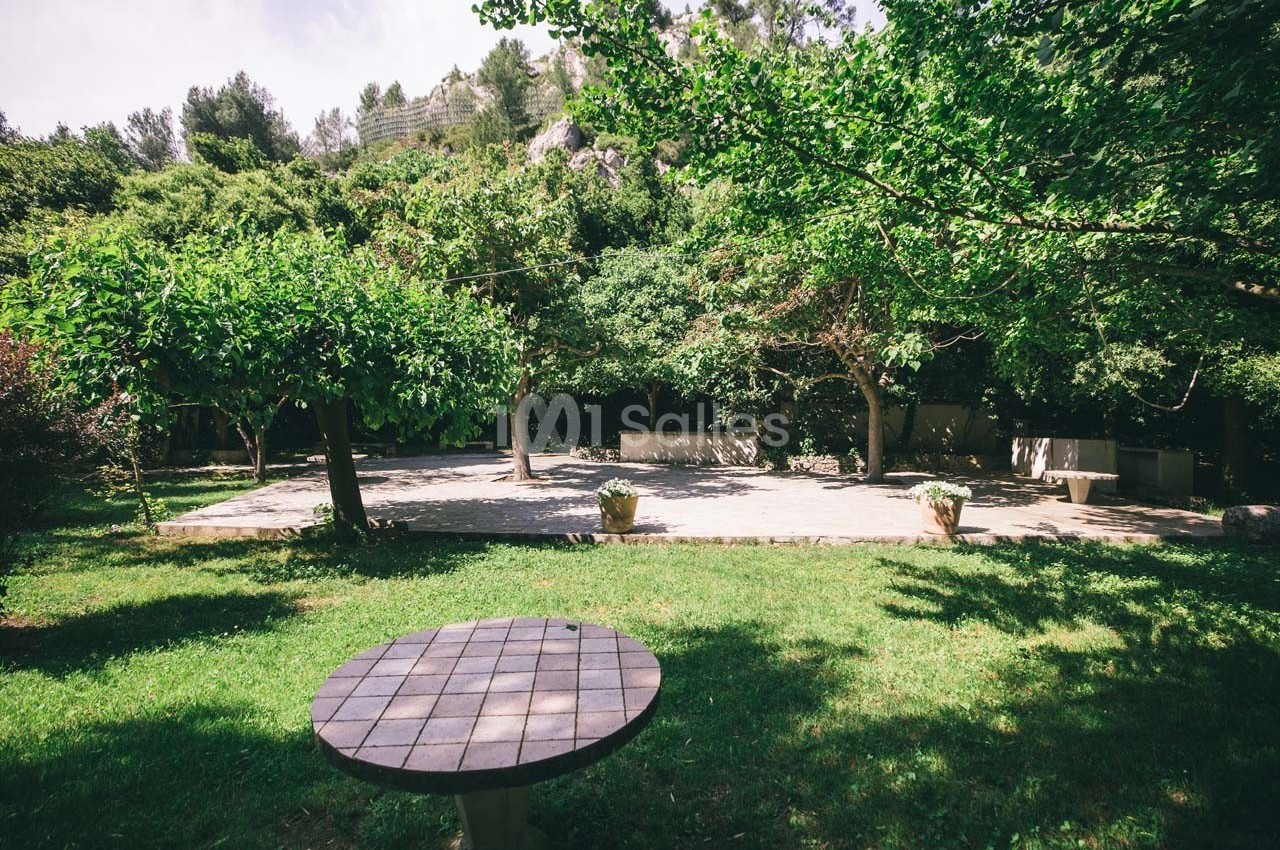 Table ronde en pierre sur une pelouse, entourée d'arbres et d'une terrasse ensoleillée en arrière-plan.