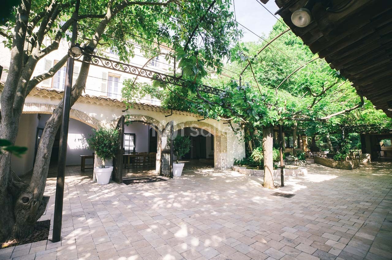 Cour intérieure pavée entourée d'arbres et de végétation, avec une façade en arches et un toit en tuiles.