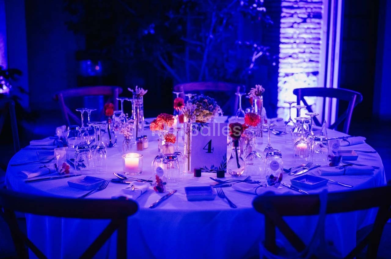 Table ronde décorée pour un dîner, avec bougies, fleurs colorées et vaisselle, éclairée par une lumière bleue.