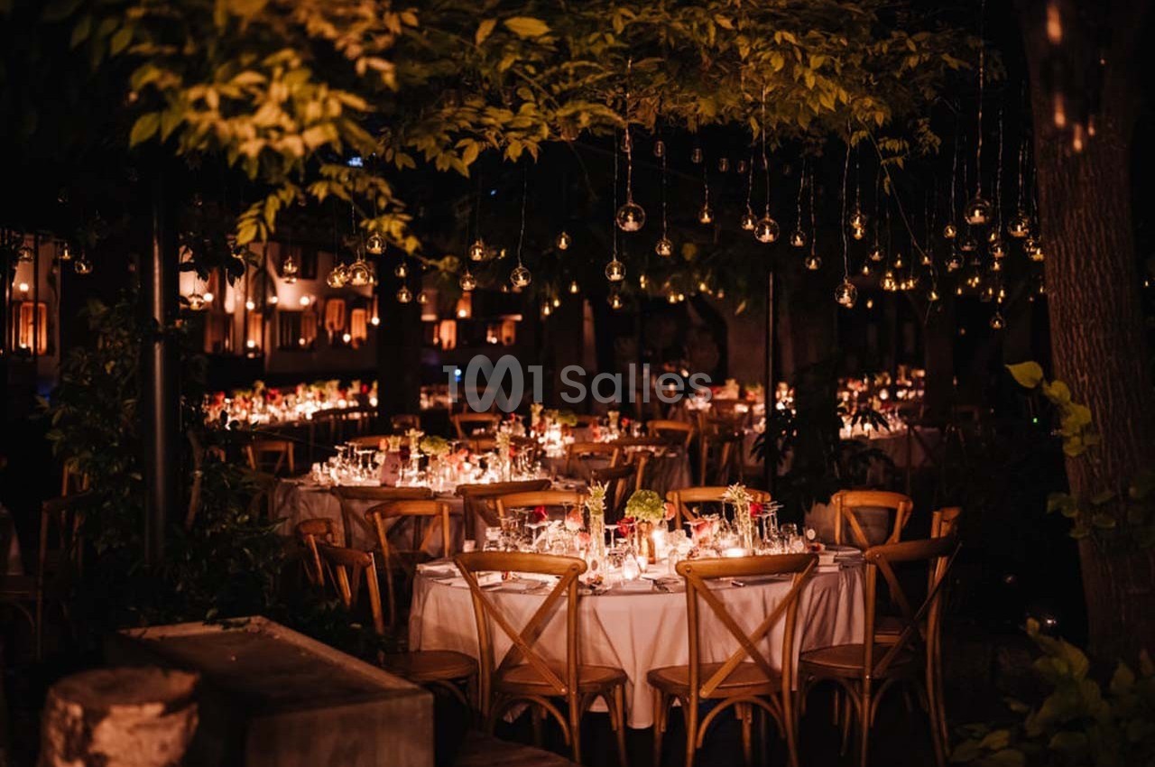 Tables décorées avec des bougies et des fleurs sous des guirlandes lumineuses dans un jardin en soirée.