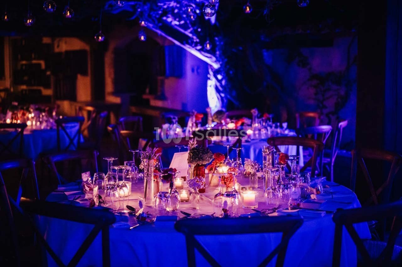 Table ronde décorée pour un dîner, éclairée par des bougies et des lumières violettes dans une ambiance nocturne.