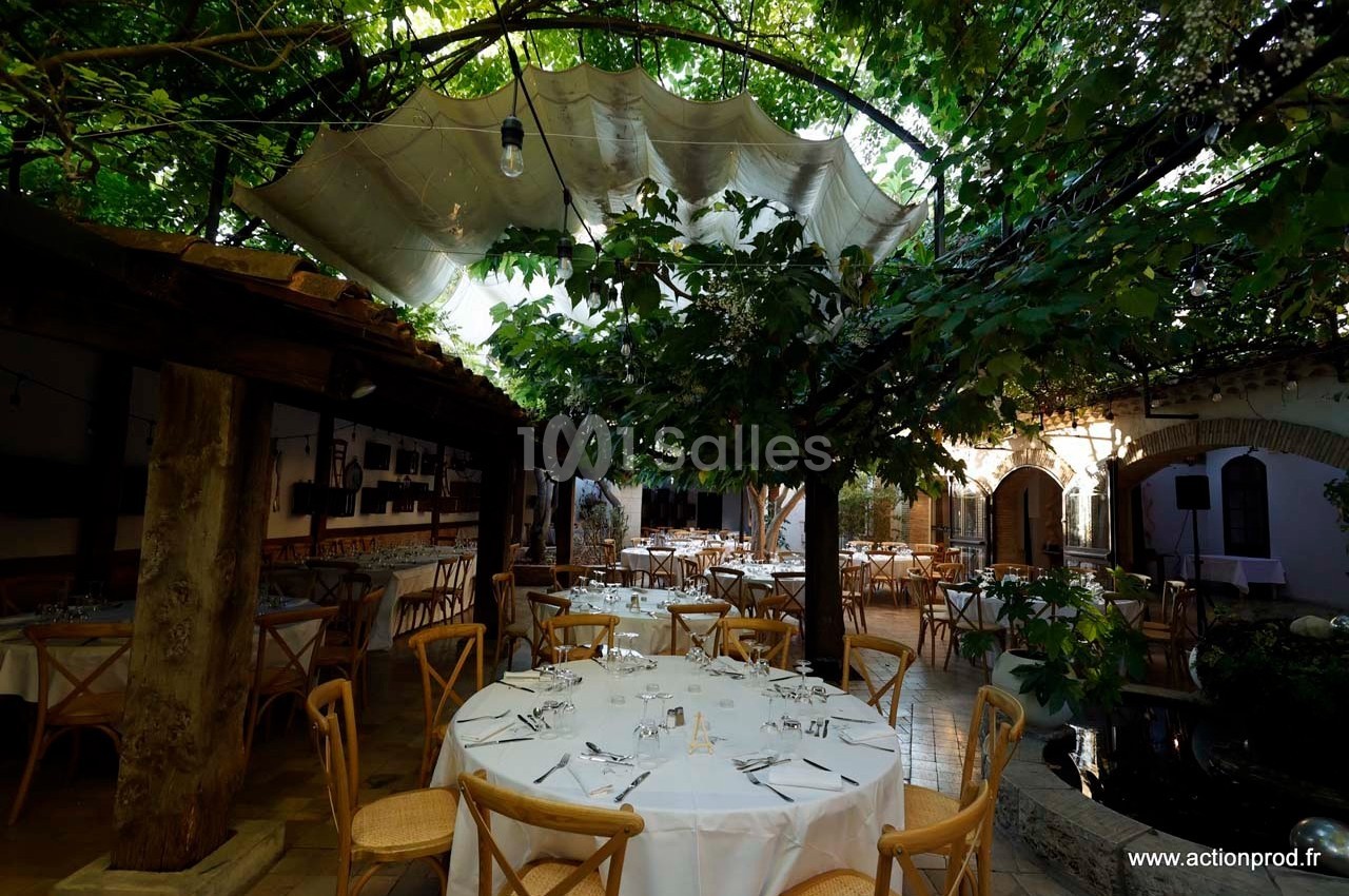 Salle de réception extérieure avec tables dressées sous une pergola ombragée par des arbres et une toile tendue.