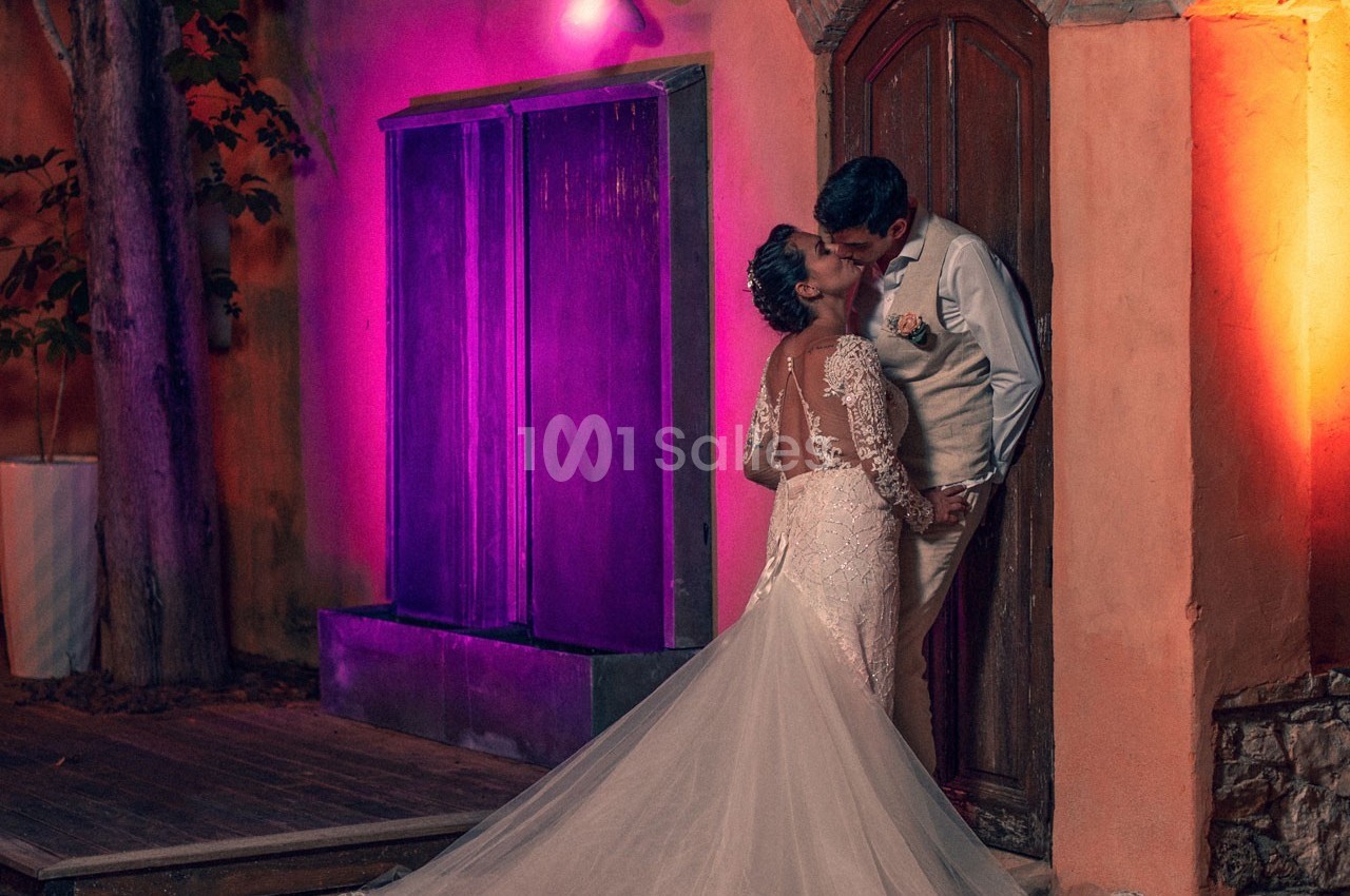 Un couple en tenue de mariage s'embrasse devant une porte en bois, éclairé par des lumières colorées.