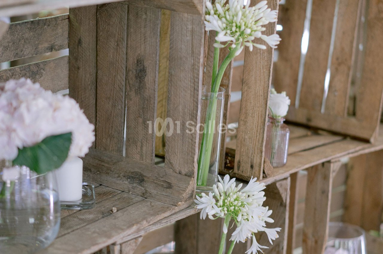 Des caisses en bois empilées contenant des vases avec des fleurs blanches et roses dans un décor rustique.