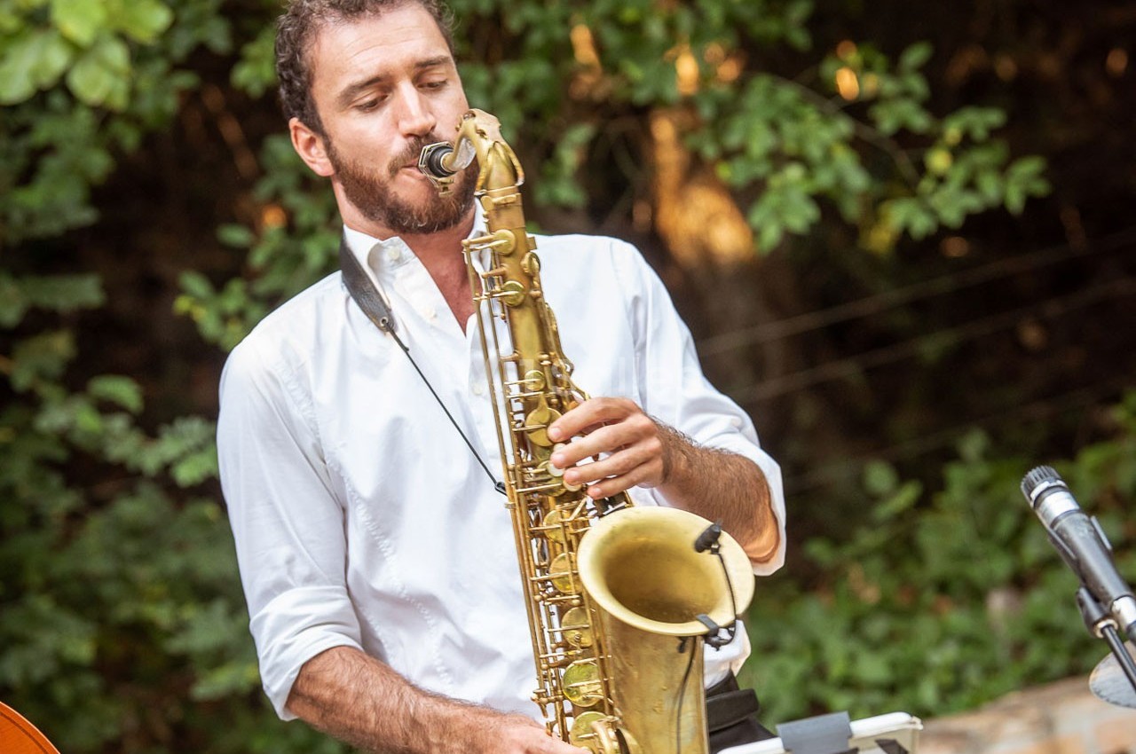 Un homme joue du saxophone en plein air, entouré de verdure.