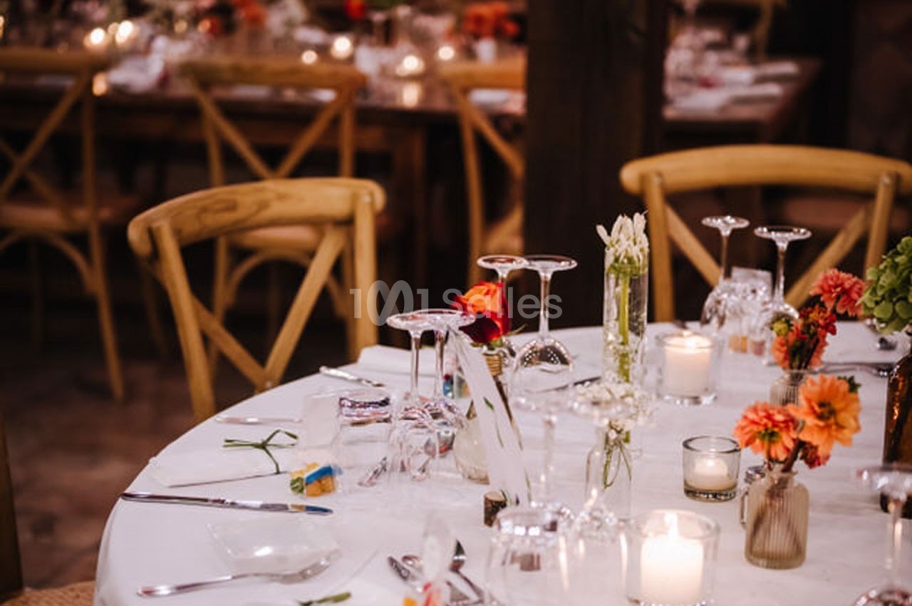 Table dressée avec nappes blanches, verres, bougies et fleurs colorées, dans une salle avec des chaises en bois.