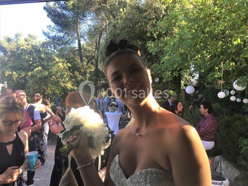 Une mariée souriante tenant un bouquet de fleurs blanches lors d'une réception en extérieur, entourée d'invités.