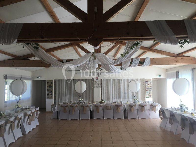 Salle de réception décorée avec des tables alignées, nappes blanches, chaises ornées et drapés suspendus au plafond.