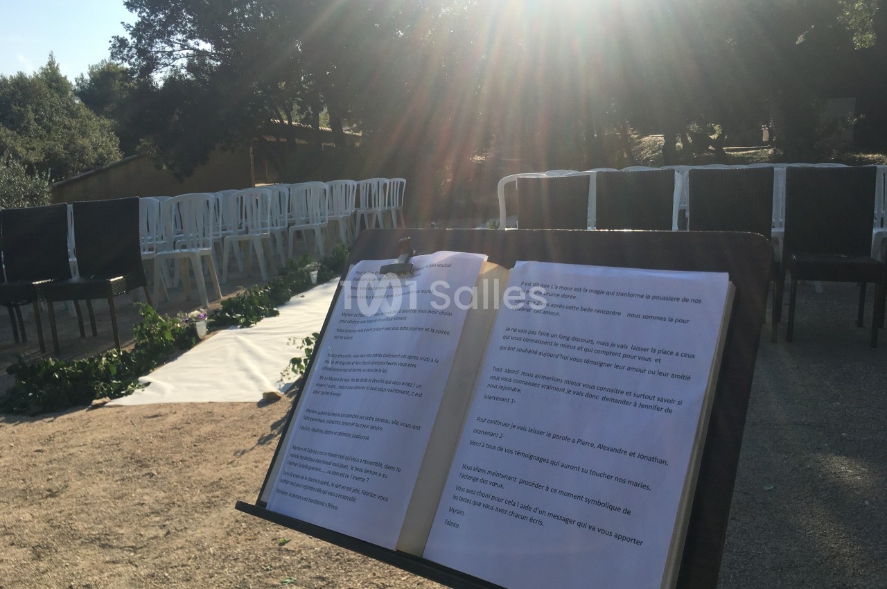 Livre ouvert sur un pupitre en plein air, devant des chaises disposées pour un événement sous un soleil éclatant.