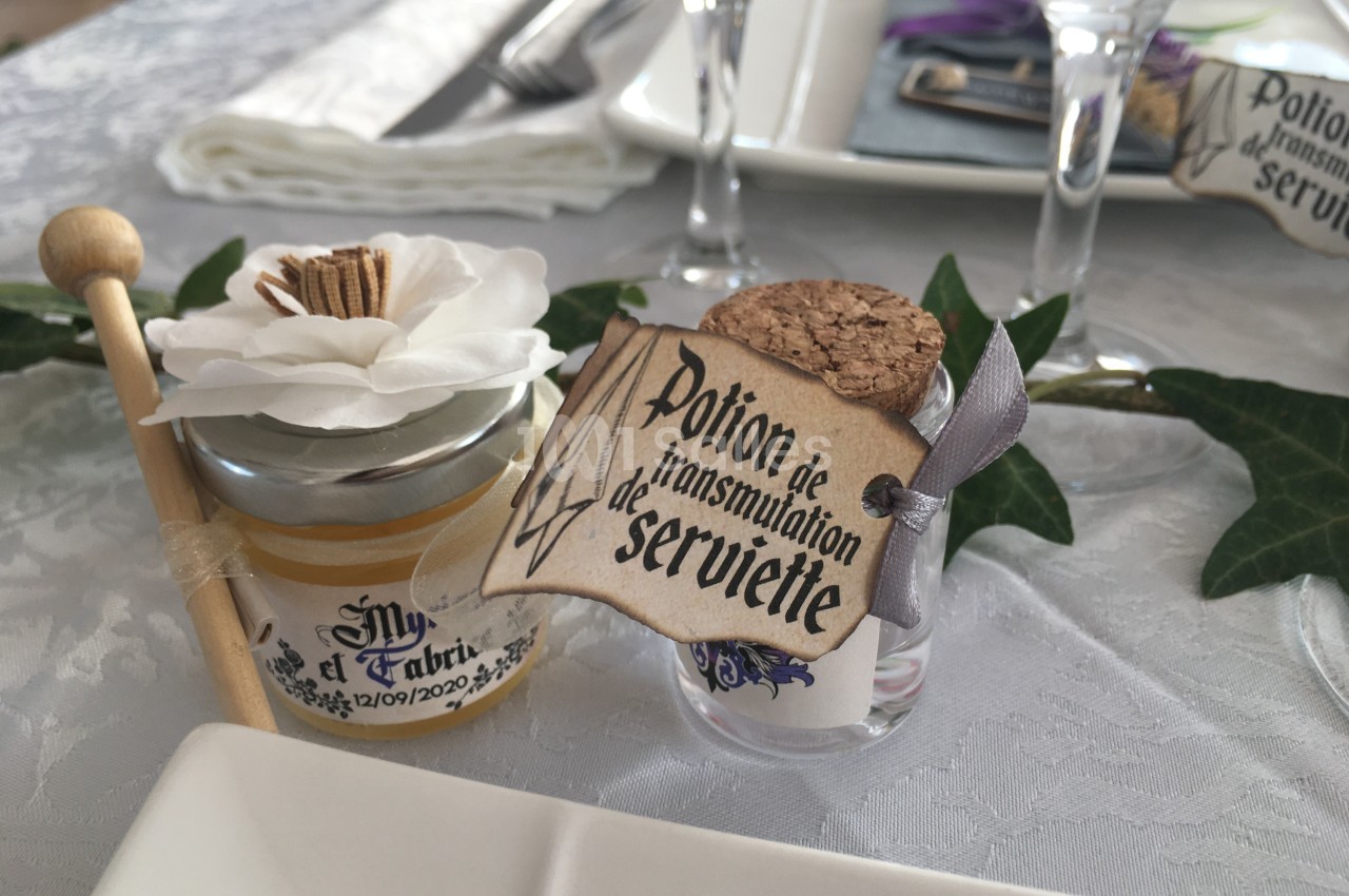 Petit pot de miel décoré avec une fleur et une fiole étiquetée ’Potion de transmutation de serviette’ sur une table dressée.
