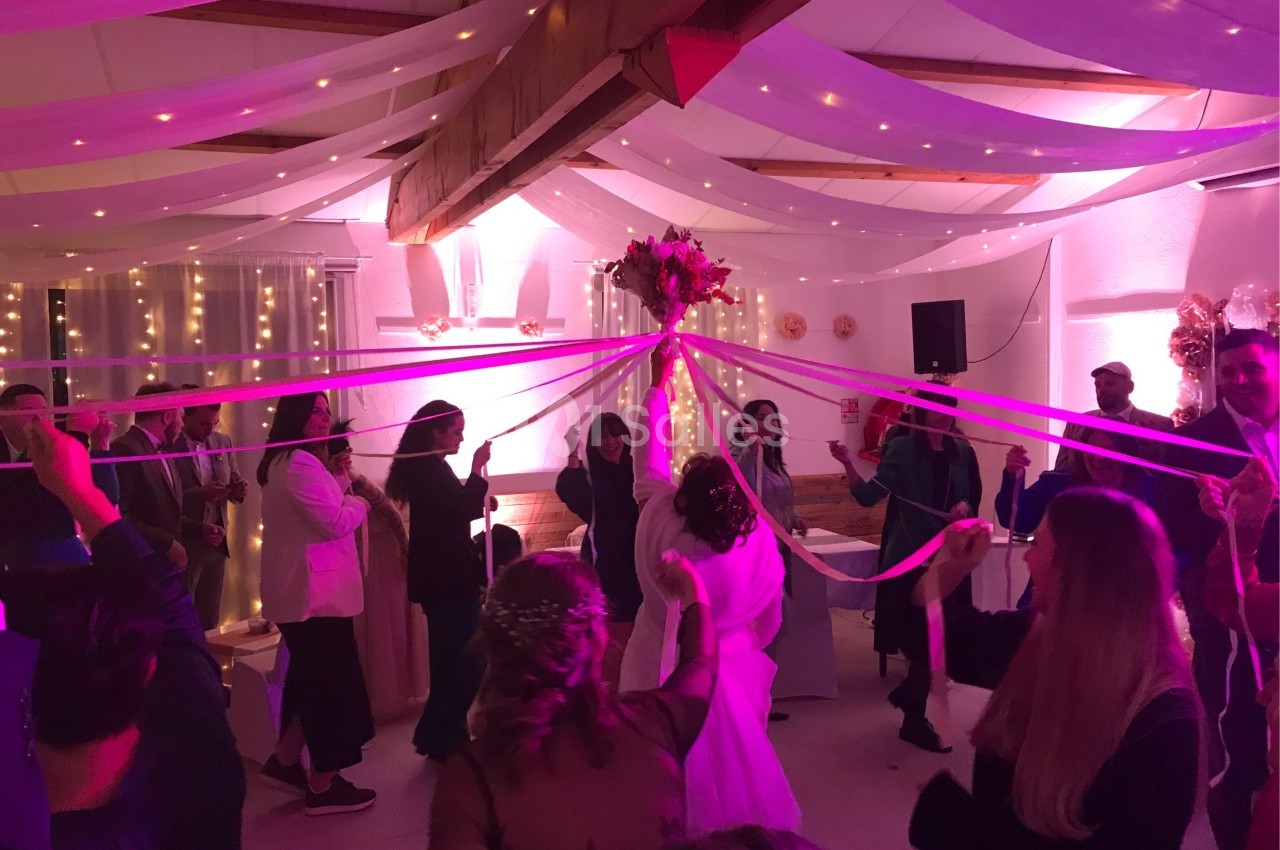 Des invités dansent autour de la mariée en tenant des rubans colorés sous un plafond décoré de guirlandes lumineuses.