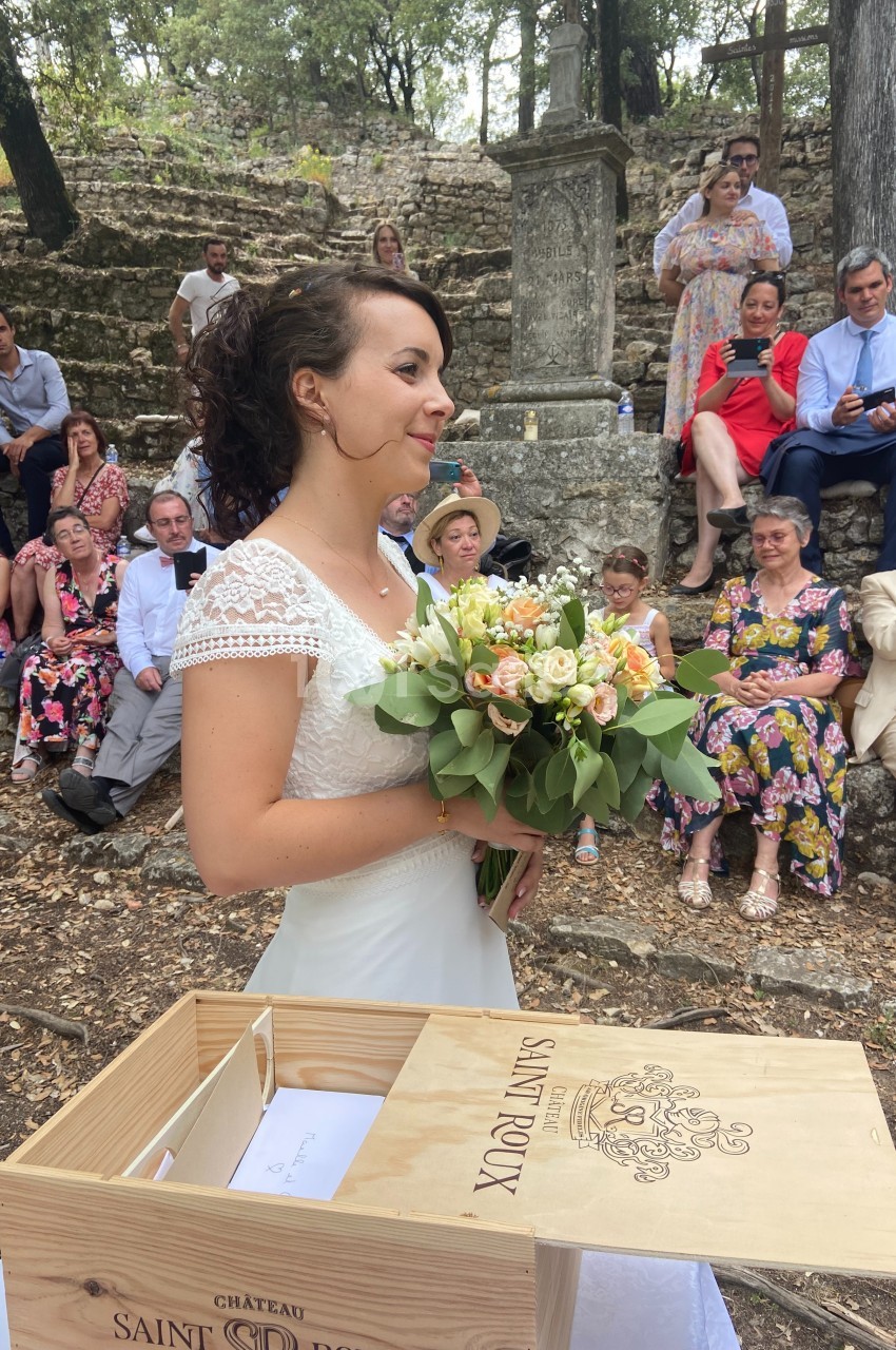 Une mariée en robe blanche tient un bouquet de fleurs devant des invités assis en plein air.