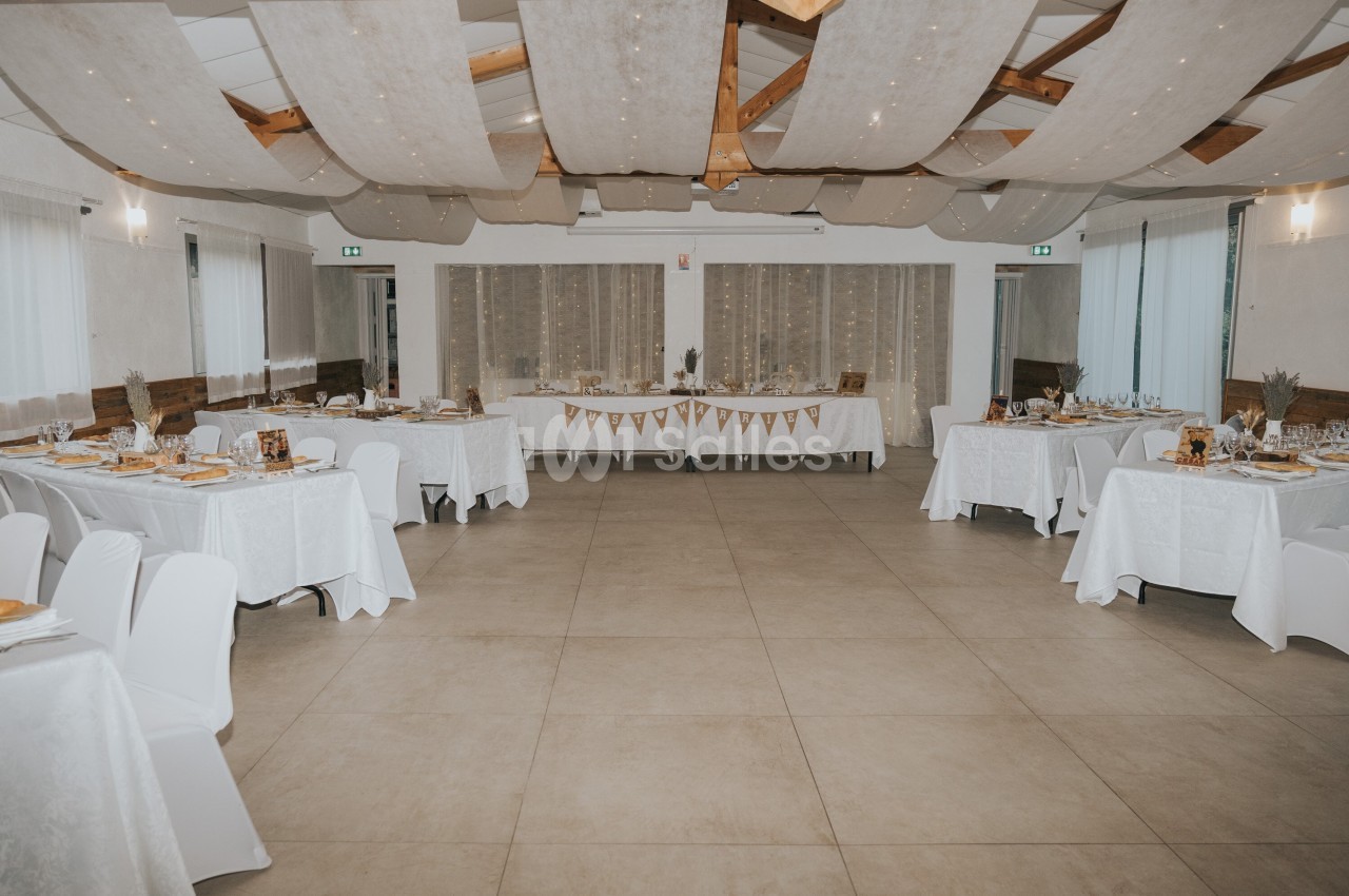Salle de réception décorée avec des tables dressées, nappes blanches et guirlandes suspendues au plafond.