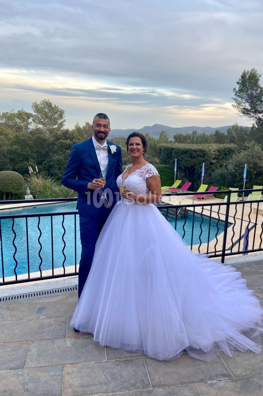 Un couple en tenue de mariage pose devant une piscine extérieure avec un paysage arboré en arrière-plan.