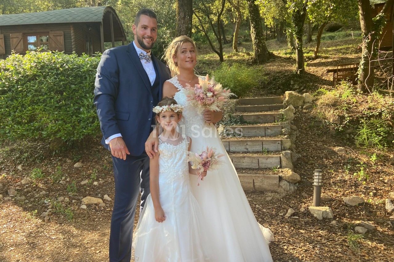 Un couple en tenue de mariage pose avec une fillette dans un jardin boisé près d'escaliers en pierre.