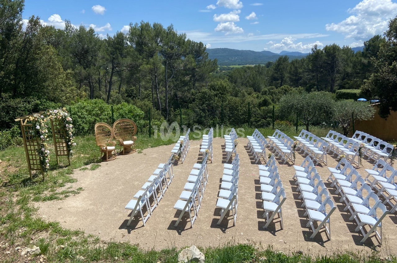Chaises blanches disposées en rangées face à une arche fleurie dans un cadre naturel avec vue sur des collines boisées.