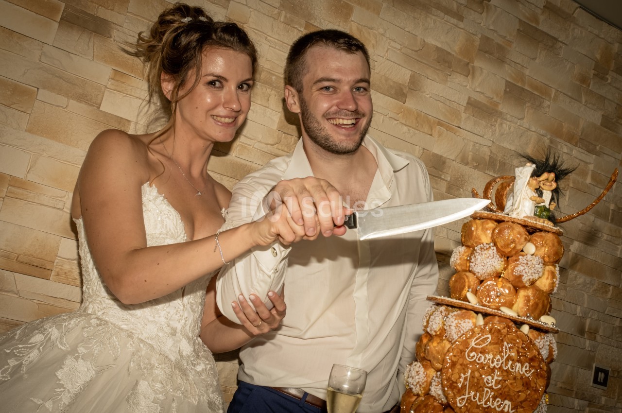 Un couple en tenue de mariage coupe une pièce montée décorée, devant un mur en pierre.