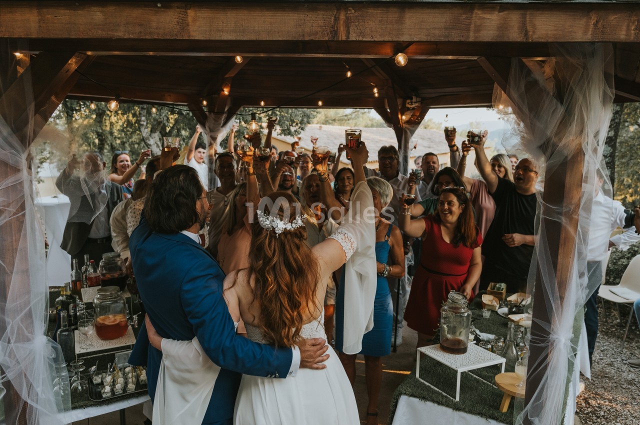 Un couple de mariés lève un verre avec des invités sous une pergola décorée, lors d'une célébration en plein air.