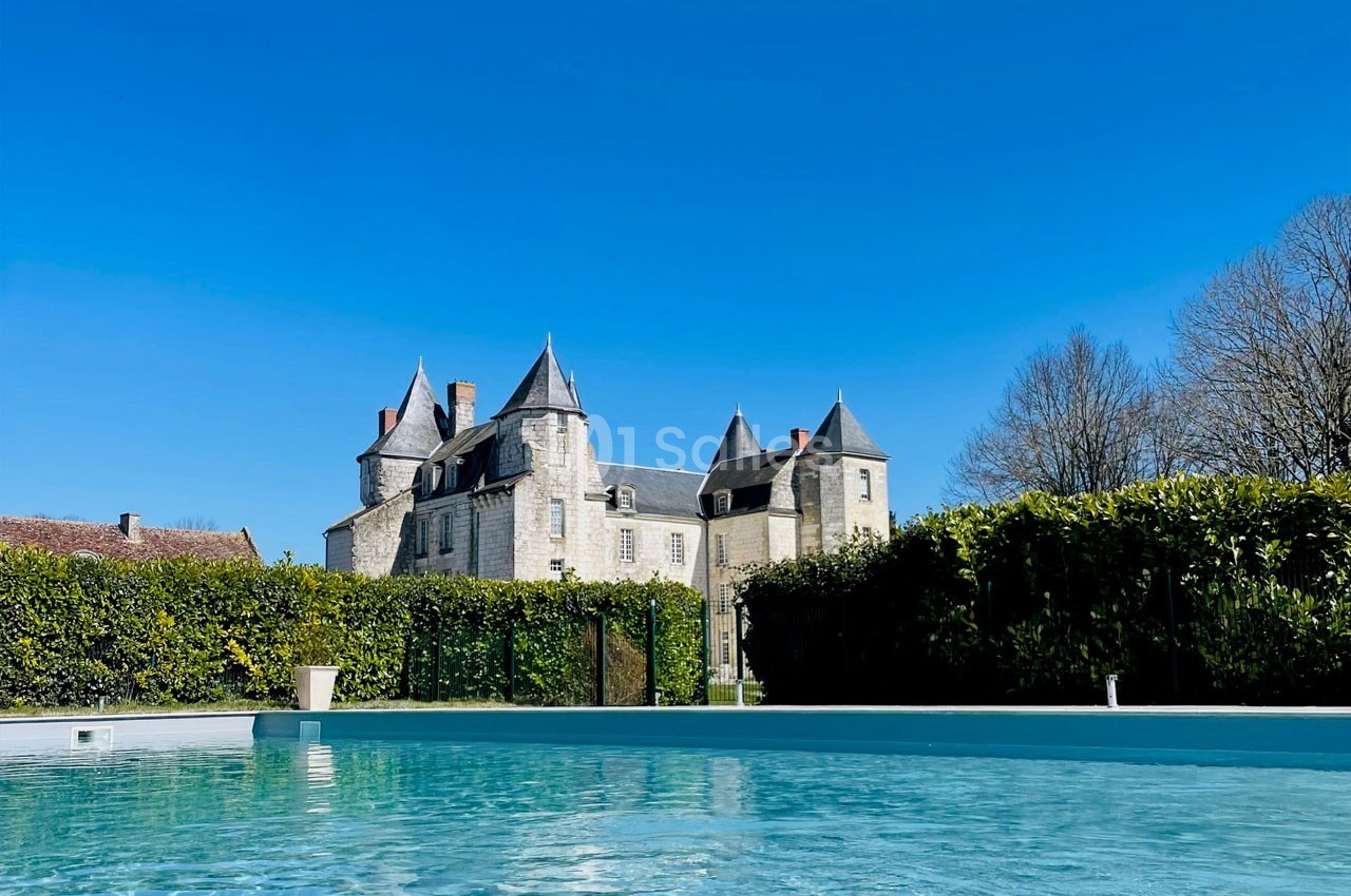 Château en pierre entouré de haies et d'arbres, avec une piscine au premier plan sous un ciel bleu clair.