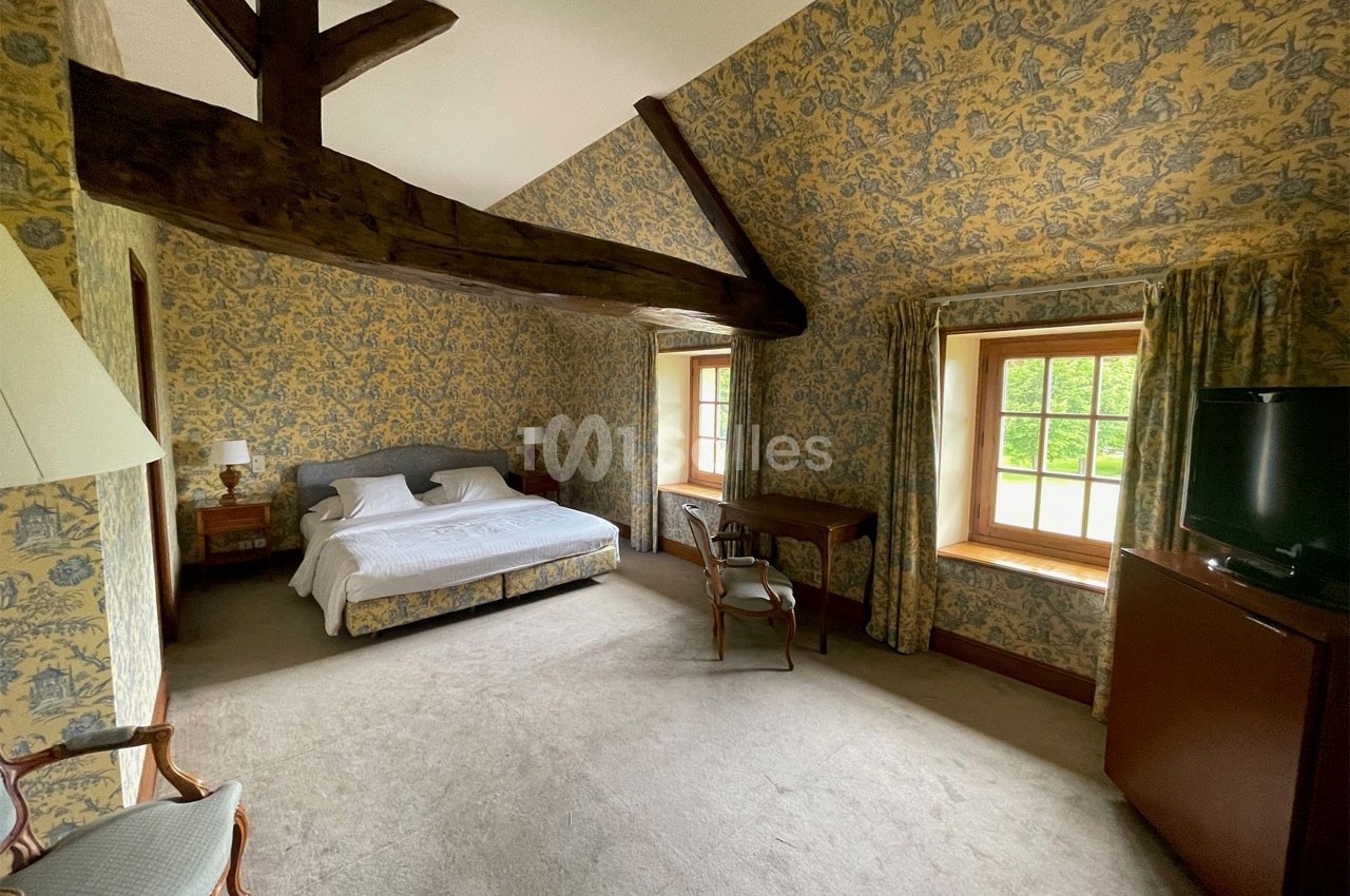 Chambre lumineuse avec lit double, poutres apparentes, murs tapissés, bureau, fauteuil et fenêtres donnant sur un jardin.