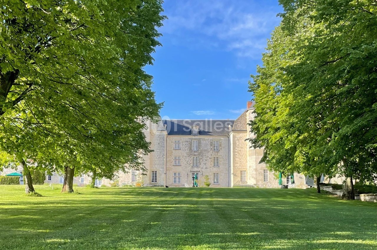 Façade d'un château en pierre entouré d'arbres et d'une pelouse verte sous un ciel bleu dégagé.