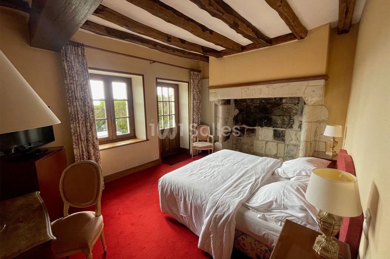 Chambre avec lit double, poutres apparentes, cheminée en pierre, moquette rouge et vue sur un jardin par une fenêtre.